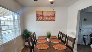 Une salle à manger avec une table en bois mise pour six personnes, entourée de chaises, un ventilateur de plafond et un tableau accroché au mur.