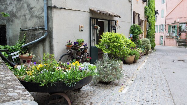 A narrow cobblestone street with a wheelbarrow of flowers, a bicycle, and potted plants along the buildings. - Villa with Independent Studio, Pool, Jacuzzi, Playground, Quiet Nature (CHATEAUVERT)
