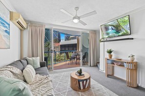 A modern living room with a sofa, a coffee table, a TV mounted on the wall, and a balcony with outdoor furniture. - Beach Haven Sea Breeze Rooftop Terrace Dual Level (Surfers Paradise)