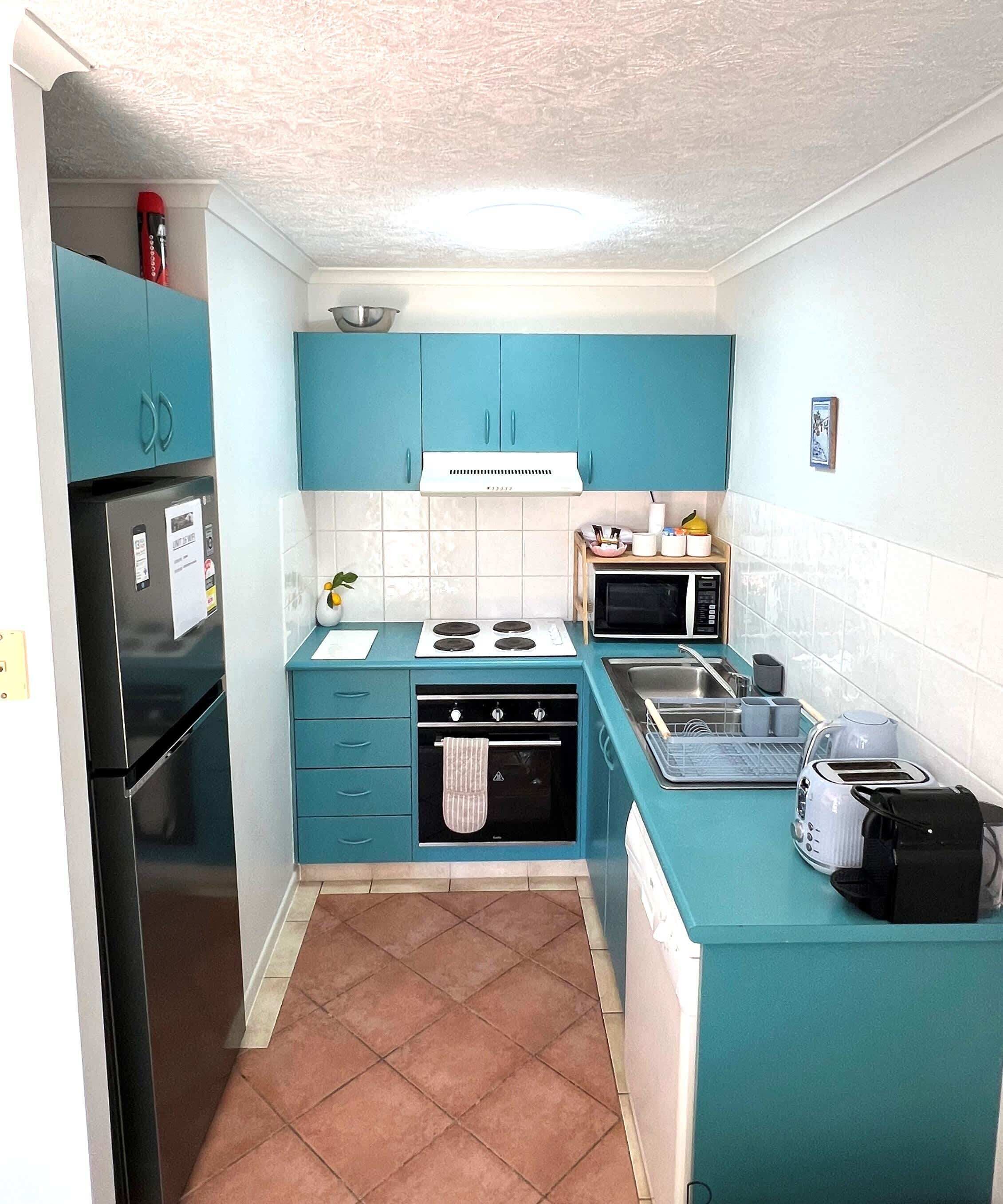 Une cuisine de taille réduite avec des meubles de couleur turquoise, un réfrigérateur noir, un four à micro-ondes, une cuisinière et un évier.