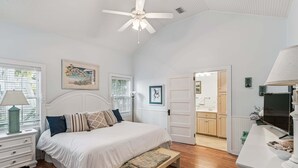 Une chambre à coucher comprenant un lit, des tables de chevet, un banc et un ventilateur de plafond, avec vue sur la salle de bain.