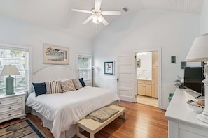 A bedroom with a bed, bedside tables, a bench, a ceiling fan, and a view into the bathroom.