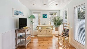 Une pièce avec un plancher en bois, un téléviseur posé sur un meuble, une armoire, une chaise et une fenêtre avec des stores.