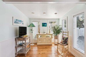 A room with a wooden floor, a television on a stand, a cabinet, a chair, and a window with blinds.
