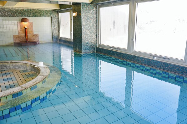 Una piscina interior con suelo y paredes de azulejos, un jacuzzi y grandes ventanales.