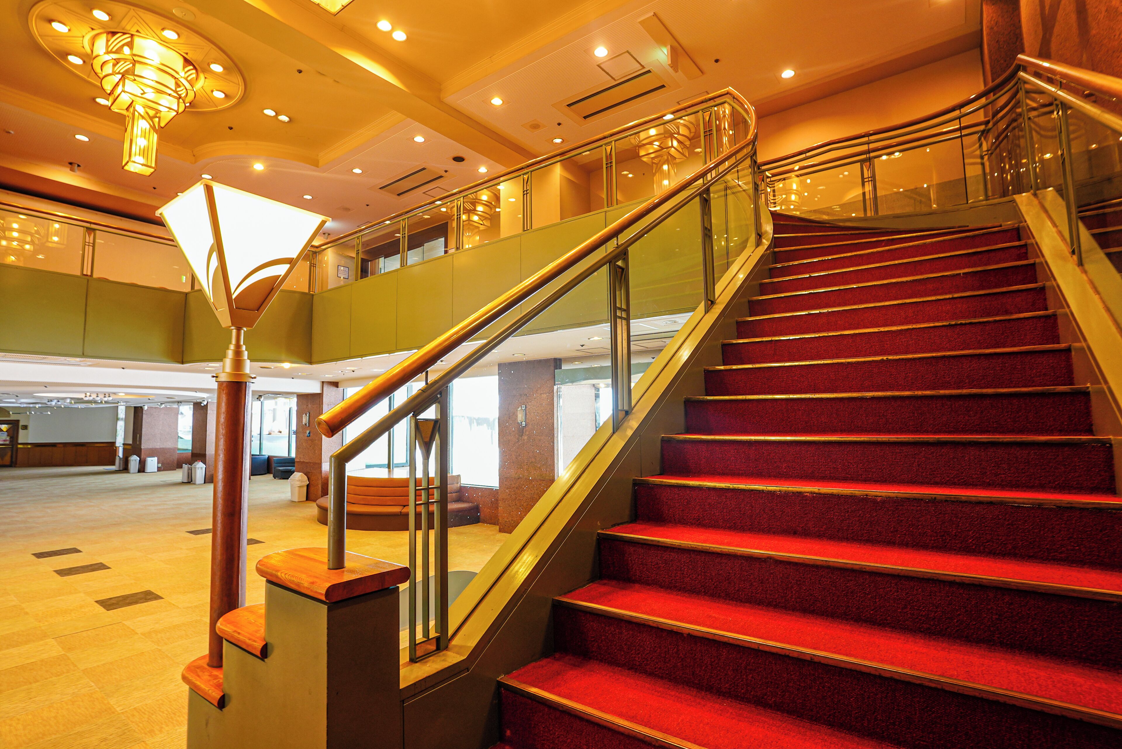 A grand staircase with red carpeted steps and a glass railing, leading to an upper level with a view of the lobby.