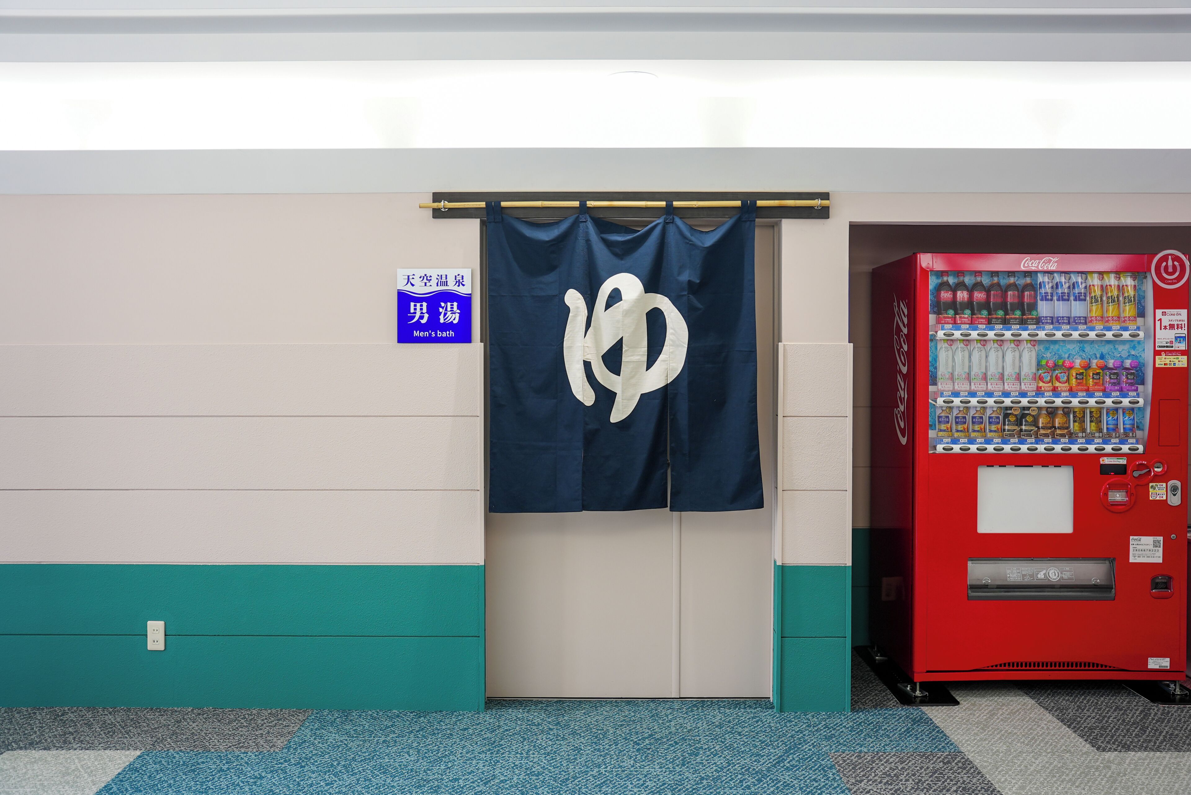 A men's bathroom with a towel hanging on a rack, a Coca-Cola vending machine, and a sign indicating the men's bath area.