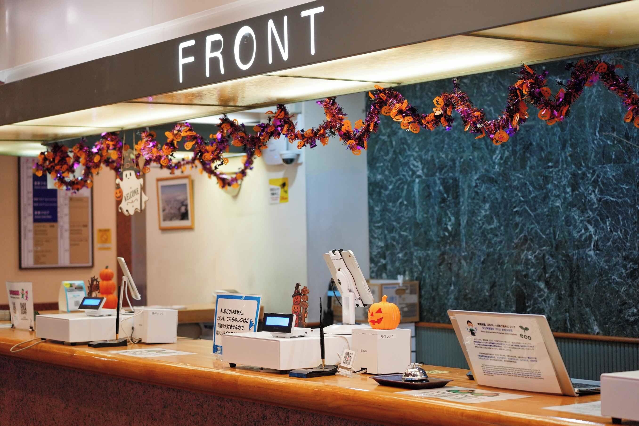 Un mostrador de recepción con un cartel que dice 'FRONT'. Hay varios dispositivos en la barra, incluyendo una impresora, una computadora y un teléfono. La pared detrás del mostrador está decorada con guirnaldas y adornos con temática de Halloween.