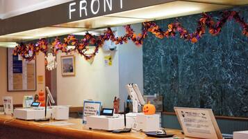 Un mostrador de recepción con un cartel que dice 'FRONT'. Hay varios dispositivos en la barra, incluyendo una impresora, una computadora y un teléfono. La pared detrás del mostrador está decorada con guirnaldas y adornos con temática de Halloween.