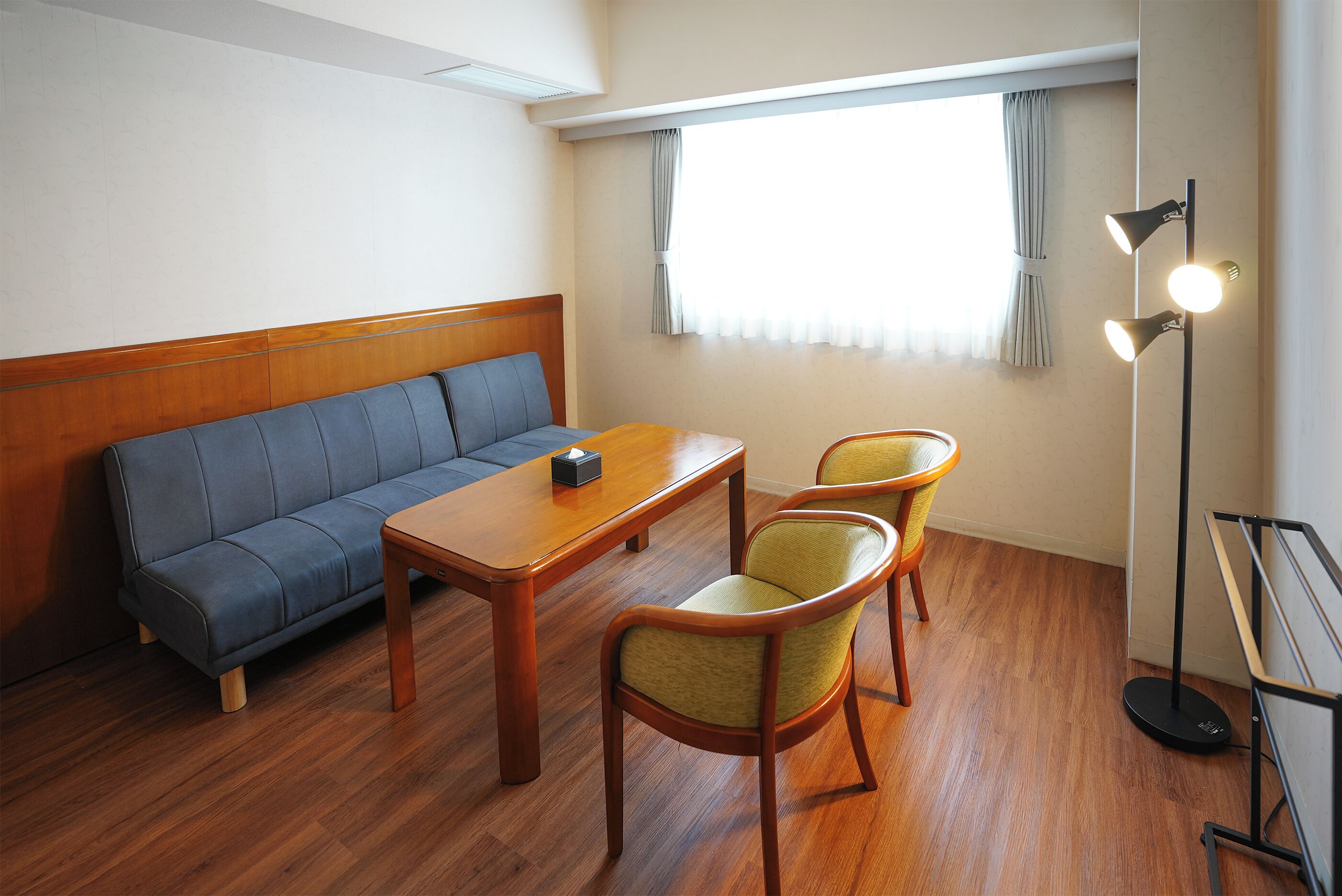A room with a wooden sofa, a small table, two chairs, a floor lamp, and a window with curtains.