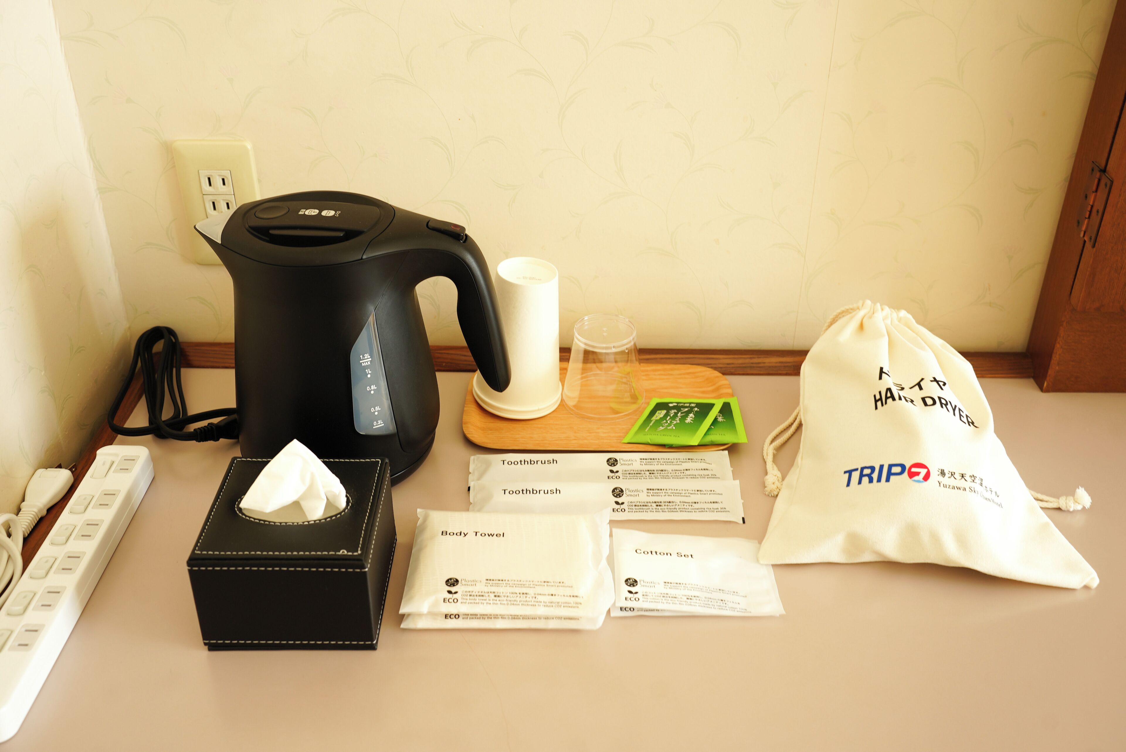 A kitchen area with a kettle, a tissue box, a glass, a tea set, and a bag of toiletries.