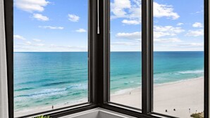 Blick aus einem Fenster auf einen Strand mit klarem, blauem Wasser und sandigem Ufer.