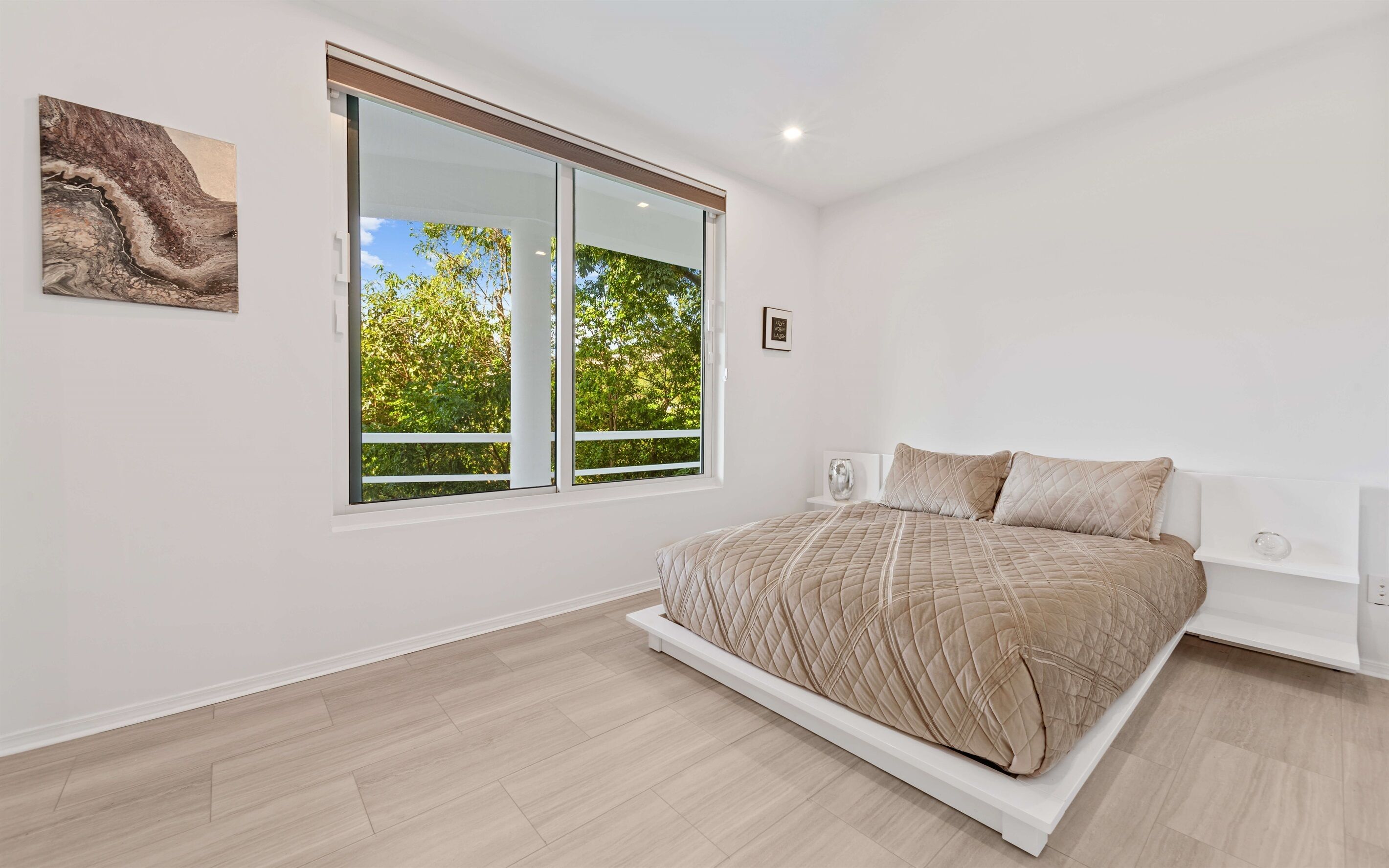 A bedroom with a large window, a bed with a quilted cover, and a piece of abstract art on the wall.