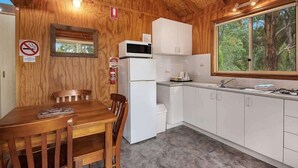 Cocina compacta con gabinetes blancos, microondas y pileta. Hay una mesa de madera con sillas. Una ventana ofrece vistas a árboles fuera.