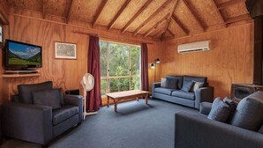 A cozy living room with wooden walls, a large window with curtains, a flat-screen TV, a sofa, a coffee table, and an air conditioning unit. - Canobolas Mountain Two-Bedroom Cabin 1 (Canobolas)
