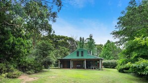 A rustic cabin with a green roof, surrounded by dense greenery and a clear sky.