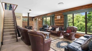 A log cabin interior with a staircase, leather sofas, a fireplace, and large windows offering a view of the outdoors.