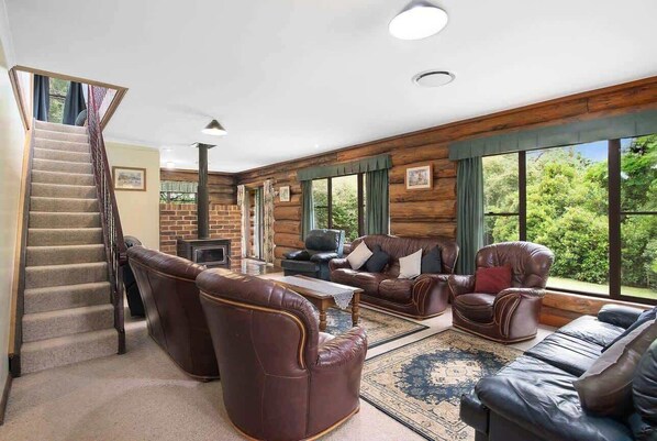 A log cabin interior with a staircase, leather sofas, a fireplace, and large windows offering a view of the outdoors.