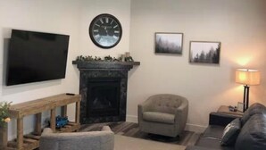 Un salon avec une cheminée, un téléviseur à écran plat, une horloge et deux tableaux encadrés accrochés au mur.