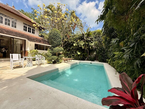 A swimming pool with a tiled deck, surrounded by lush greenery and a building with a tiled roof. - Villa White Orchid (Seminyak)