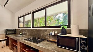 A compact kitchen with a built-in gas stove, a microwave, and a sink. There is a window above the sink, and a trash bin is visible to the left. - Villa White Orchid (Seminyak)