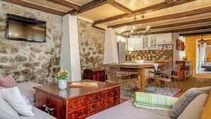 Een gezellige woonkamer met een stenen muur, houten balken, een televisie, een eethoek met een houten tafel en stoelen, en een keuken op de achtergrond.
