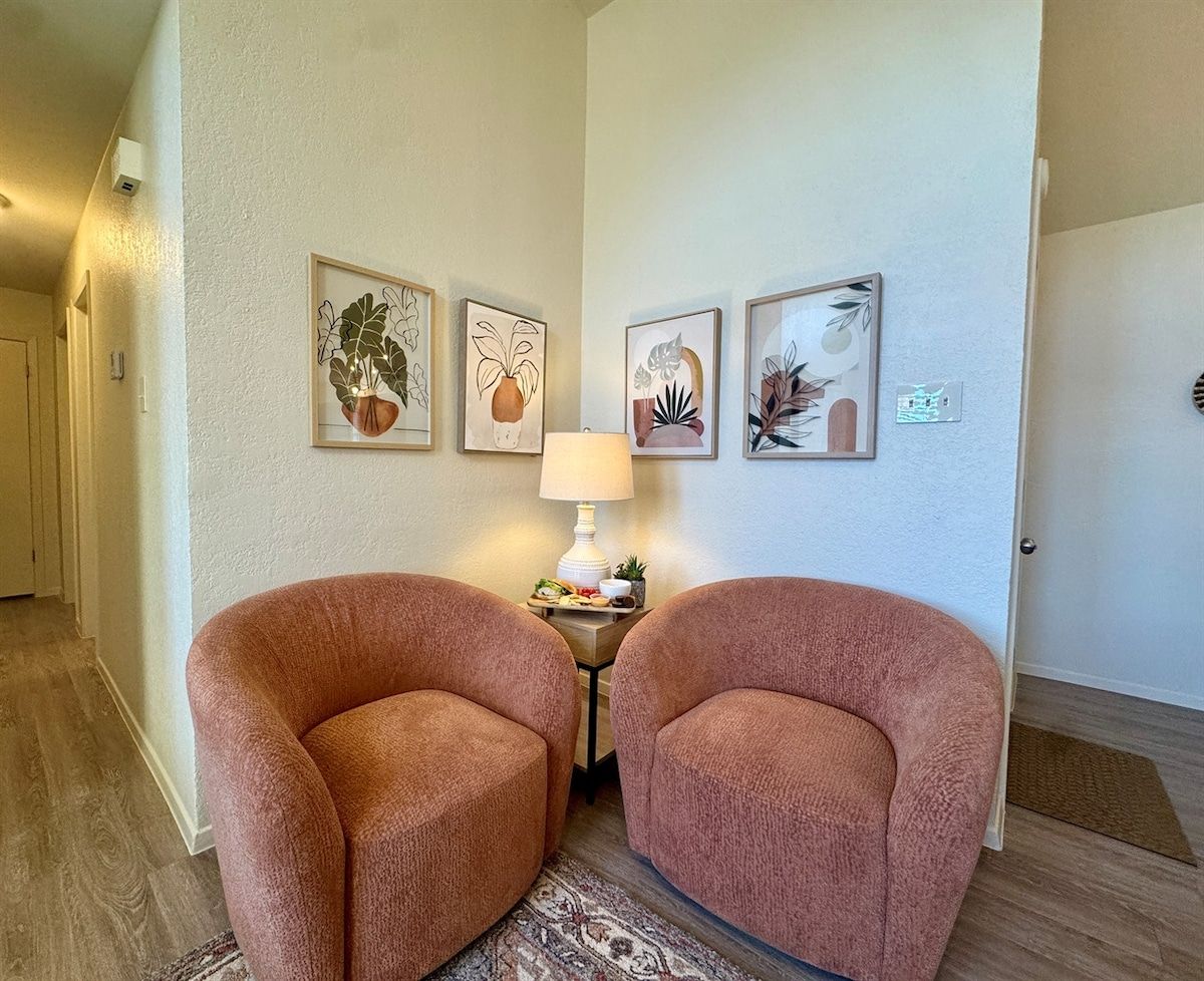 A room with two plush armchairs, a small table with a lamp and decorative items, and three framed botanical prints on the wall.