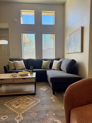 A living room with a sectional sofa, a coffee table, and a patterned rug. There are two windows with blinds, a door, and a wall-mounted artwork. - Baylor House Stylish Abilene Getaway (Abilene)