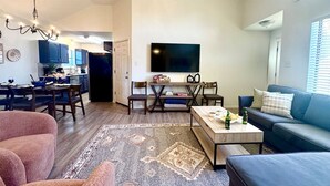 Ein Wohnzimmer mit Sofa, Couchtisch und Teppich. Es gibt einen Essbereich mit Tisch und Stühlen. Ein Fernseher ist an der Wand befestigt.