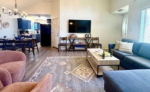 A living room with a sofa, a coffee table, and a rug. There is a dining area with a table and chairs. A TV is mounted on the wall. - Baylor House - Stylish Abilene Getaway (Abilene)