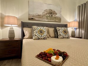 A hotel room with a bed, two bedside lamps, a nightstand, a fruit platter, and a painting on the wall. - Baylor House - Stylish Abilene Getaway (Abilene)