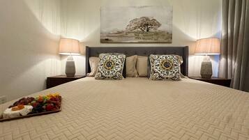 A hotel room with a large bed, two bedside lamps, a painting on the wall, and a fruit platter on the bed.
