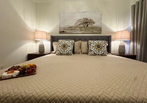 A hotel room with a large bed, two bedside lamps, a painting on the wall, and a fruit platter on the bed. - Baylor House Stylish Abilene Getaway (Abilene)