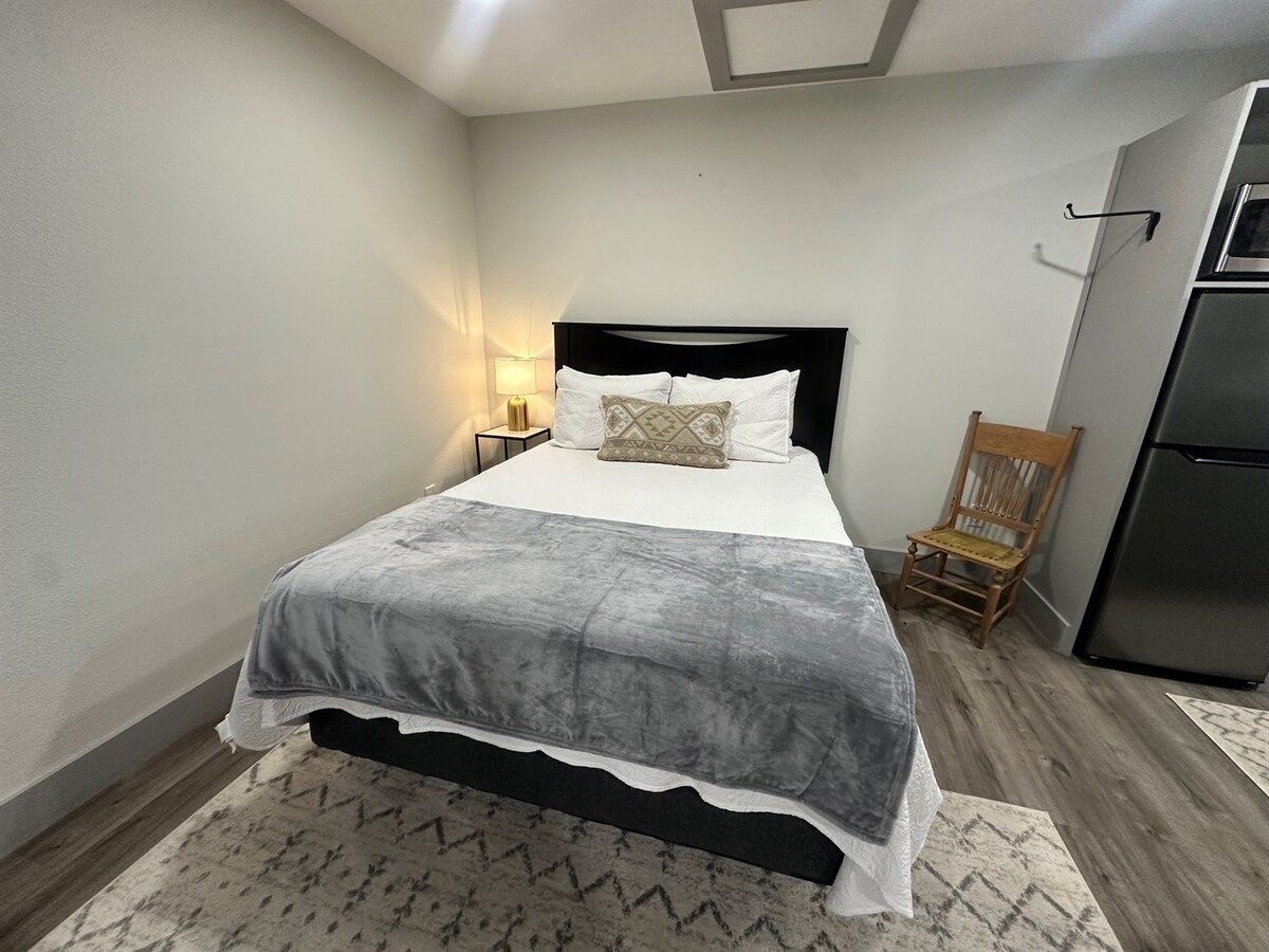 A bedroom with a bed, a nightstand, a lamp, a chair, and a refrigerator.