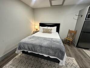 Quarto com cama, mesa de cabeceira, luminária, cadeira e geladeira.