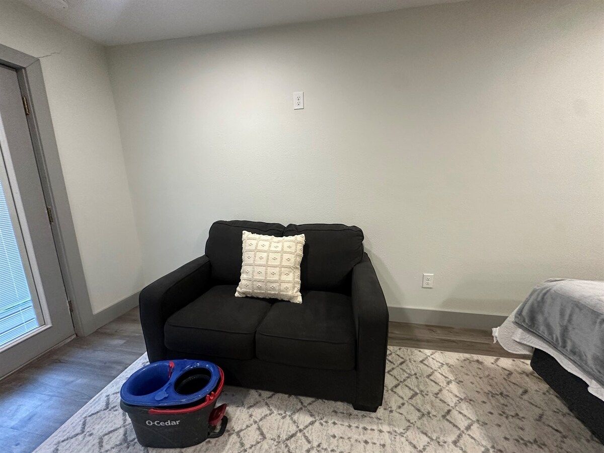 A room with a black sofa, a patterned rug, a blue and red pet bowl, and a bed with a gray blanket.