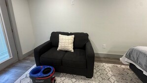A room with a black sofa, a patterned rug, a blue and red pet bowl, and a bed with a gray blanket. - Carriage House - Modern Downtown Studio (Abilene)