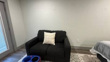 A room with a black sofa, a patterned rug, a blue and red pet bowl, and a bed with a gray blanket.
