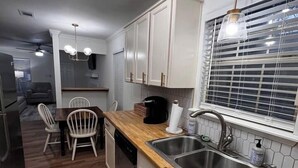 Una cocina moderna con encimeras de madera, pileta de acero inoxidable y armarios blancos. Hay un comedor con mesa y sillas, y una ventana con persianas.