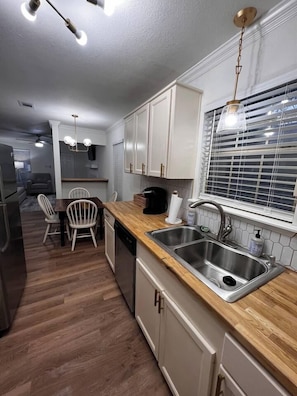 Una cocina moderna con encimeras de madera, pileta de acero inoxidable y armarios blancos. Hay un comedor con mesa y sillas, y una ventana con persianas.