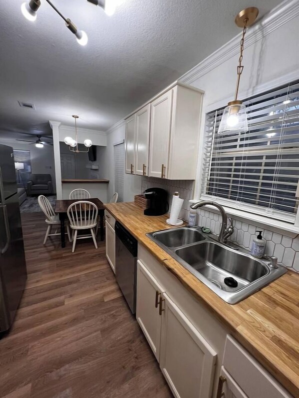 A modern kitchen with wooden countertops, stainless steel sink, and white cabinets. There is a dining area with a wooden table and chairs, and a window with blinds.