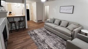 Una sala de estar moderna con un sofá gris, una mesita redonda pequeña y una alfombra gris. Se observa una zona de cocina con una barra y dos taburetes. Se ve un ventilador de techo y un cartel enmarcado en la pared.