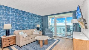 Habitación de hotel moderna con papel tapiz de un velero azul, un sofá, una mesa de centro redonda, una silla, un televisor y un balcón con vista al mar.