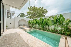 Un espace piscine extérieur moderne, doté d’une piscine à eau claire, d’un coin salon avec des coussins et d’une végétation tropicale luxuriante.