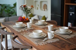 Une table de salle à manger en bois, dressée avec des bols, des assiettes et des tasses blancs, et un set de table placé en dessous.