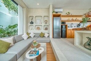 Una sala moderna con un sofá gris, una mesa de centro y una cocina con refrigerador y estantes de madera.