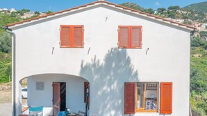 Un edificio blanco con dos ventanas y una puerta, un pequeño patio con mesa y sillas, y vistas a una colina.