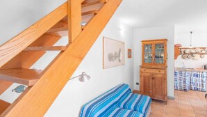 Una escalera con barandillas de madera, un sofá azul a rayas, un mueble de madera con puertas de cristal y una zona de cocina con ollas colgantes.