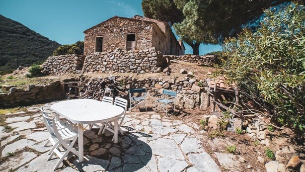 Un edificio de piedra con un ventanal pequeño y una puerta, rodeado de un patio de piedra con una mesa y sillas.