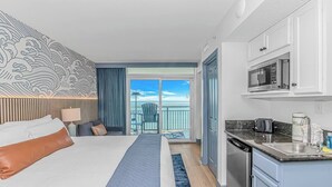 A hotel room with a bed, a kitchenette, a view of the ocean, and a balcony with a chair. - Oceanfront 2BR Retreat | 17th Floor + Balconies (Myrtle Beach)